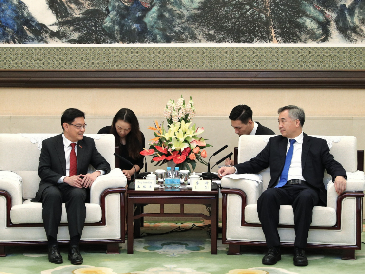 Two men in suits seated in armchairs, facing each other, with assistants, flowers, and microphones in between.